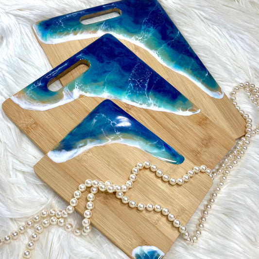Three bamboo serving boards with a blue and white ocean wave design, displayed on a white furry surface, with pearls scattered around.