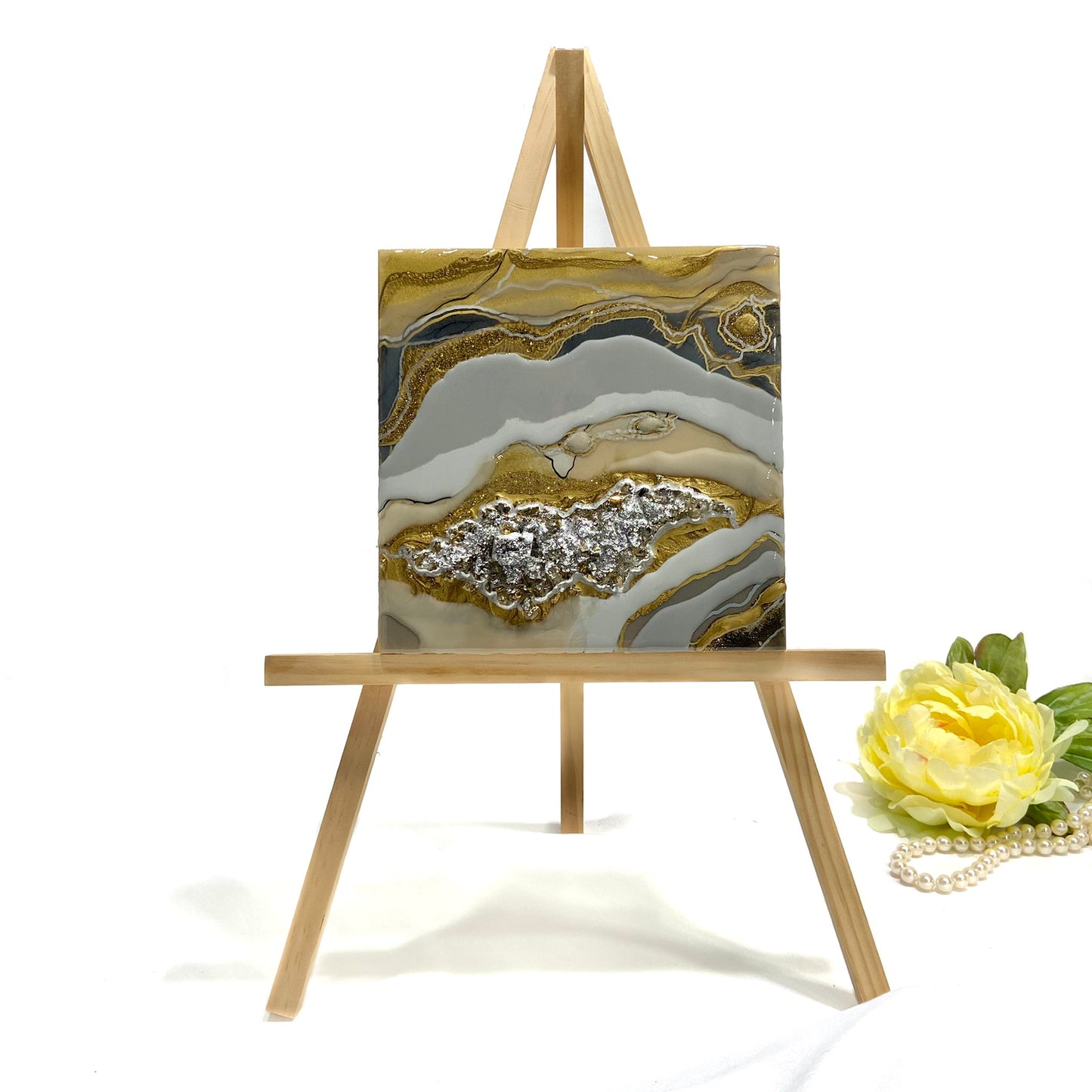 A wall art piece featuring a textured resin mix with gold and silver tones, and a central pyrite stone embellishment, presented on a birch wood panel, with a wooden frame and a decorative flower beside it.