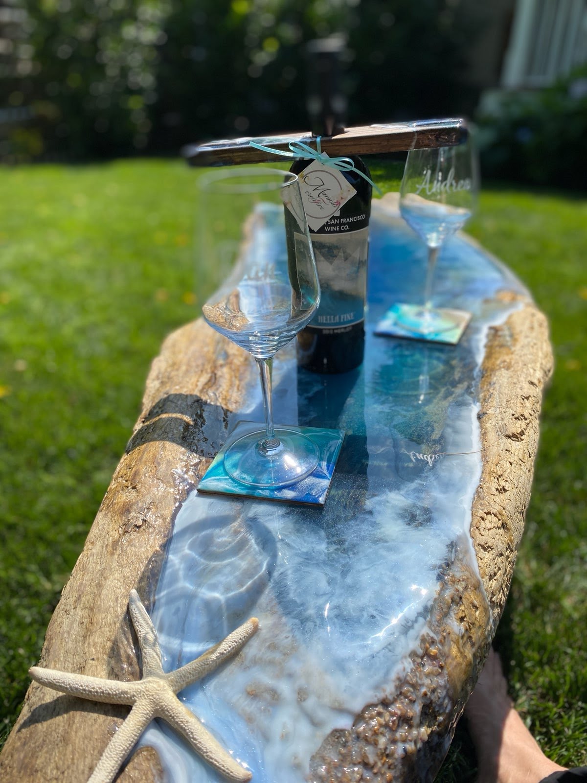 Beach theme table with sea star ornament, bottle of wine and glasses resting on ocean them coasters in a grass outdoor setting 